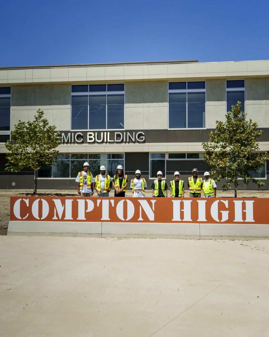 The team in hard hats in front of the Compton High sign and the new academic building