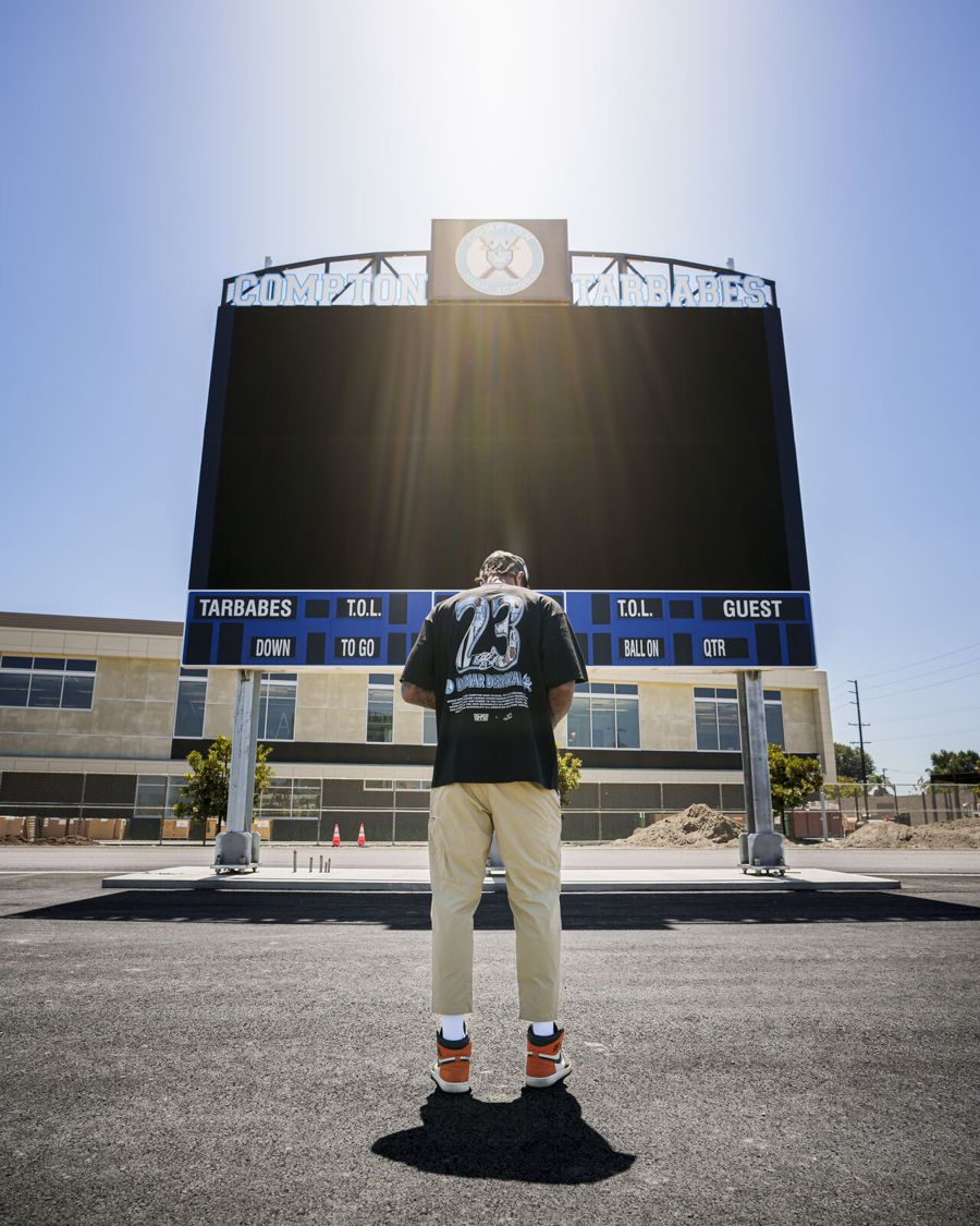 A man in a Game Changers x DeMar DeRozan shirt looking up at the scoreboard at Compton High School